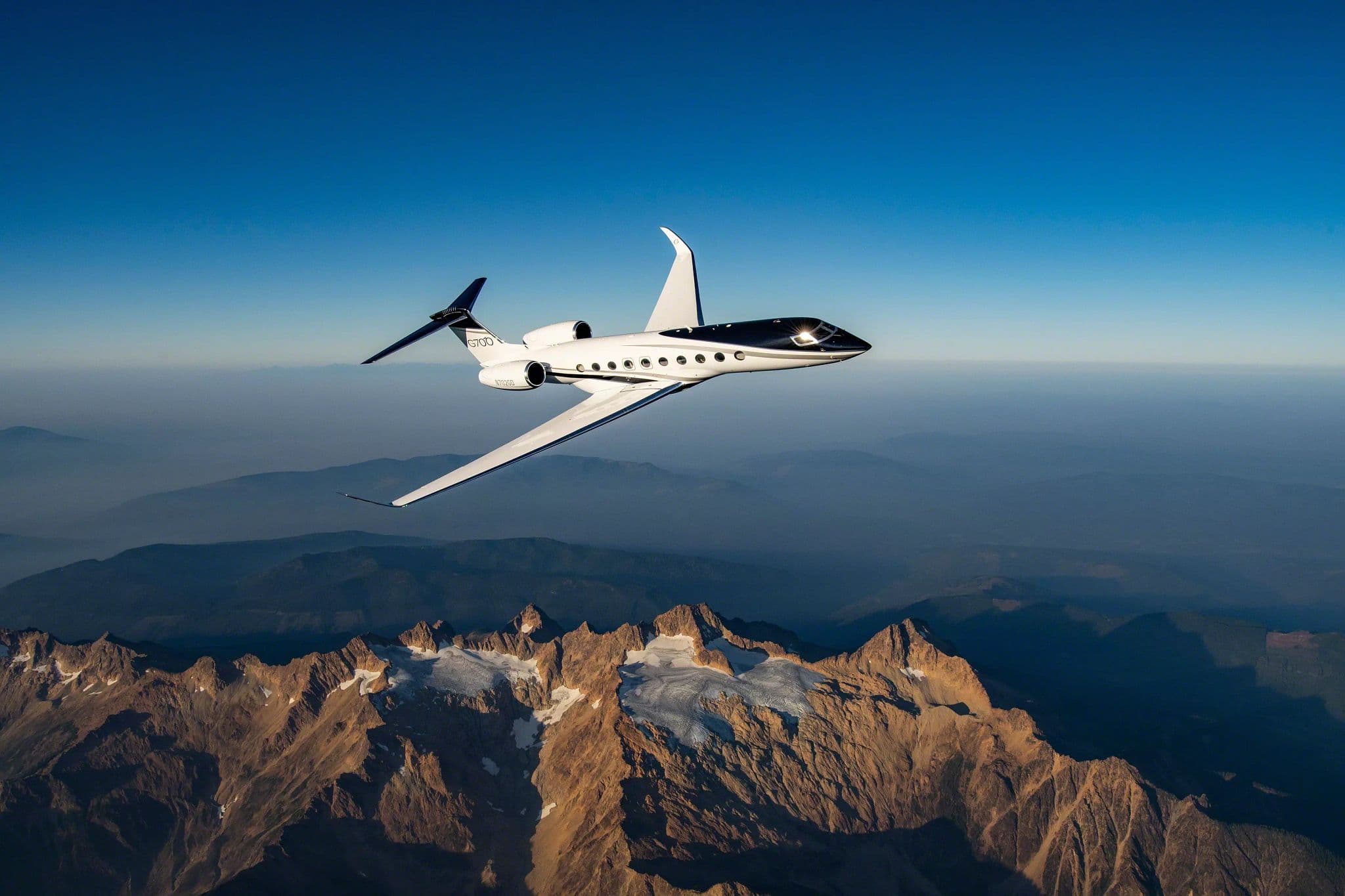 White Gulfstream G700 banking above a rugged, snow-streaked mountain range beneath a clear blue sky.